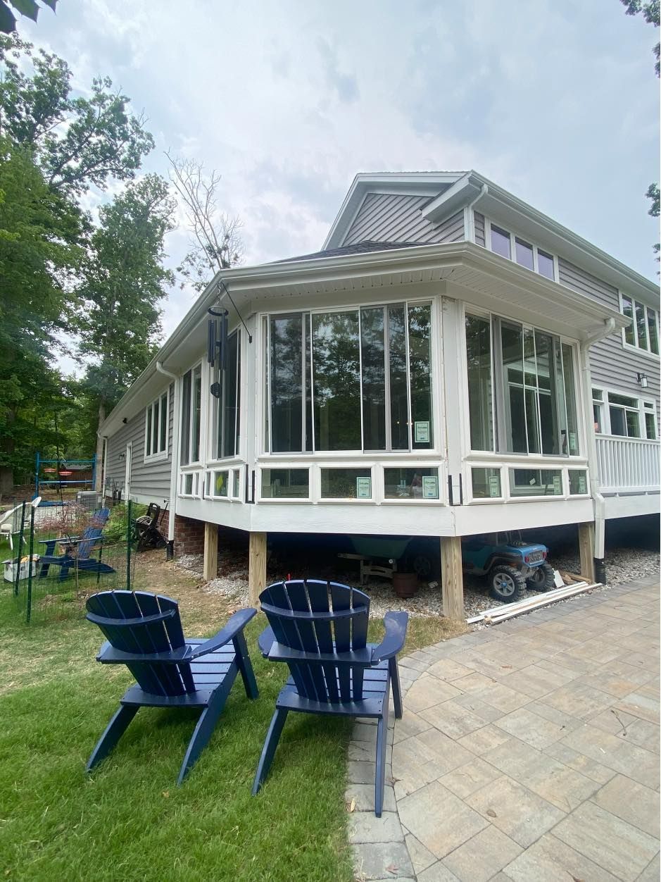 Two blue Adirondack chairs face a sunroom with large windows, on a deck of a house.