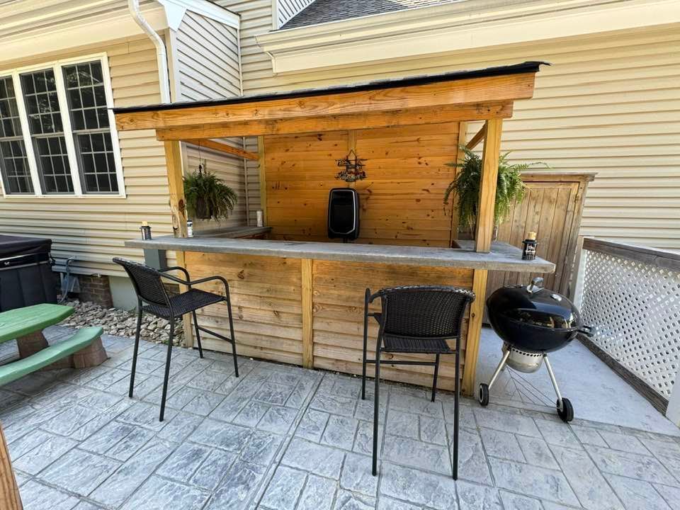 Outdoor wooden bar with stools, grill, and plants on a patio.