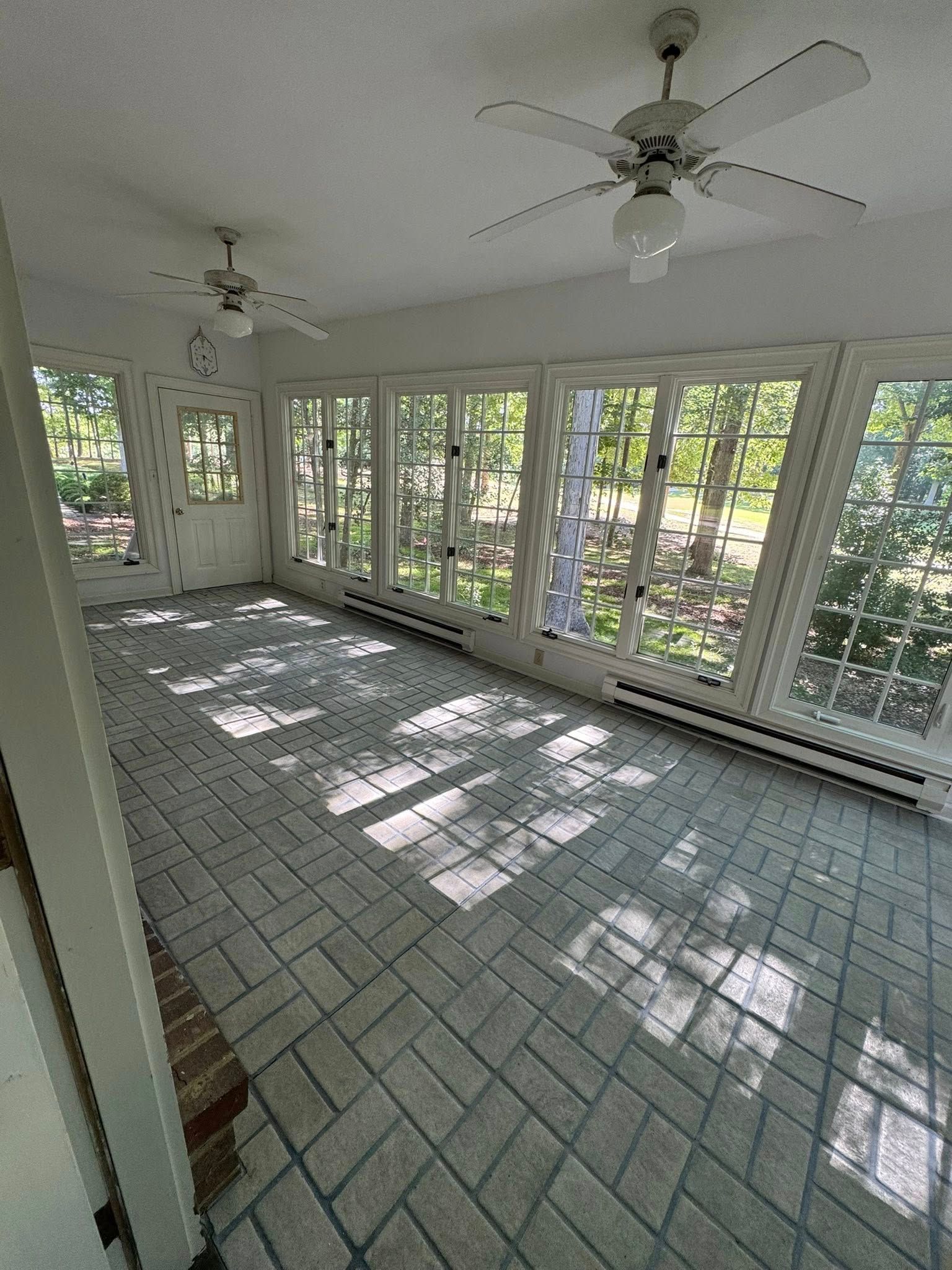 Sunroom with brick-patterned floor, white trim, and multiple windows overlooking a wooded area. Two ceiling fans.