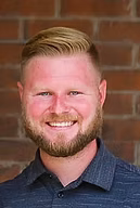 Man with a blonde, brushed-up hairstyle and beard smiles in front of a brick wall.