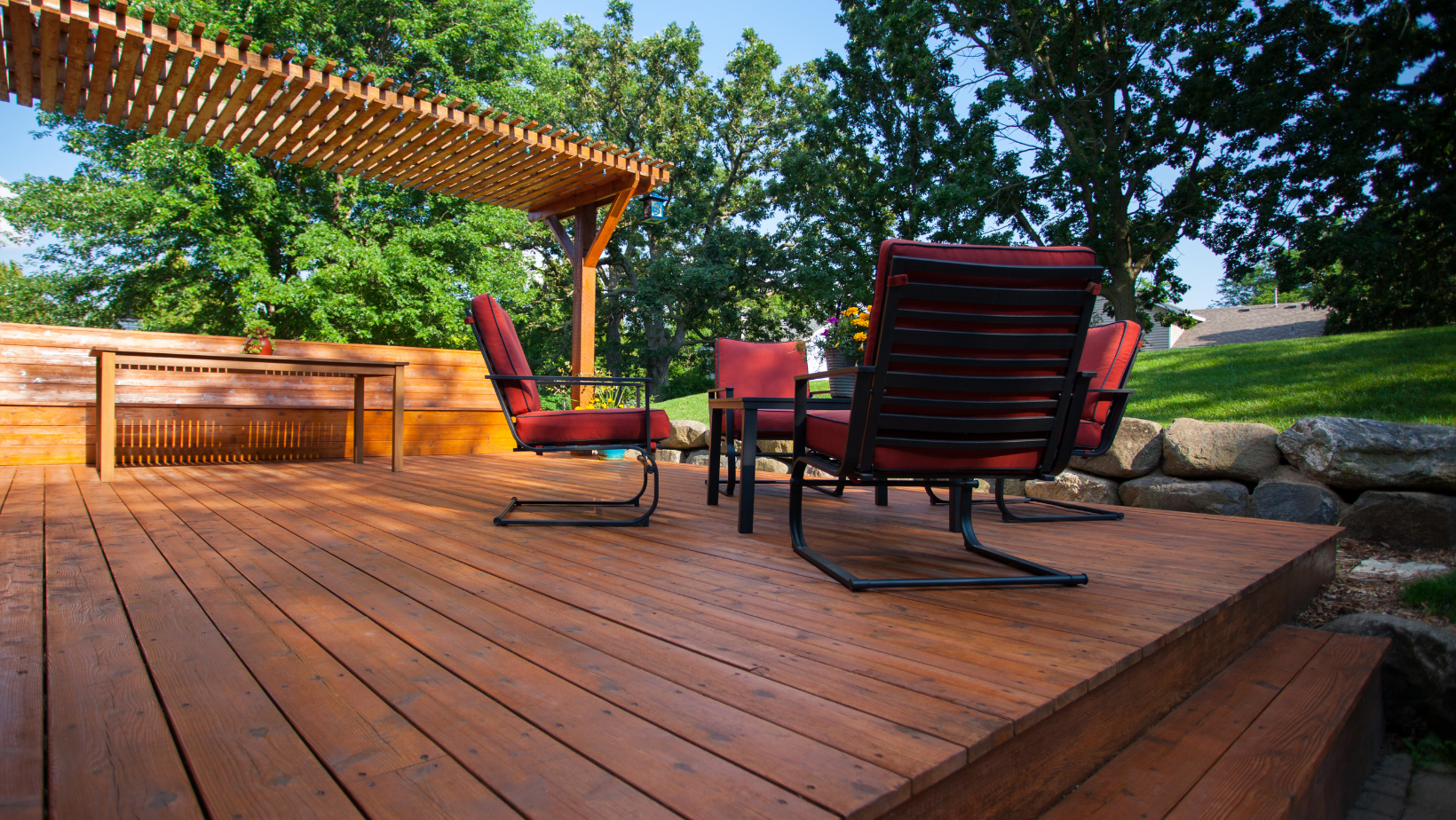 Wooden deck with red chairs and a pergola, trees in the background, sunny outdoor setting.