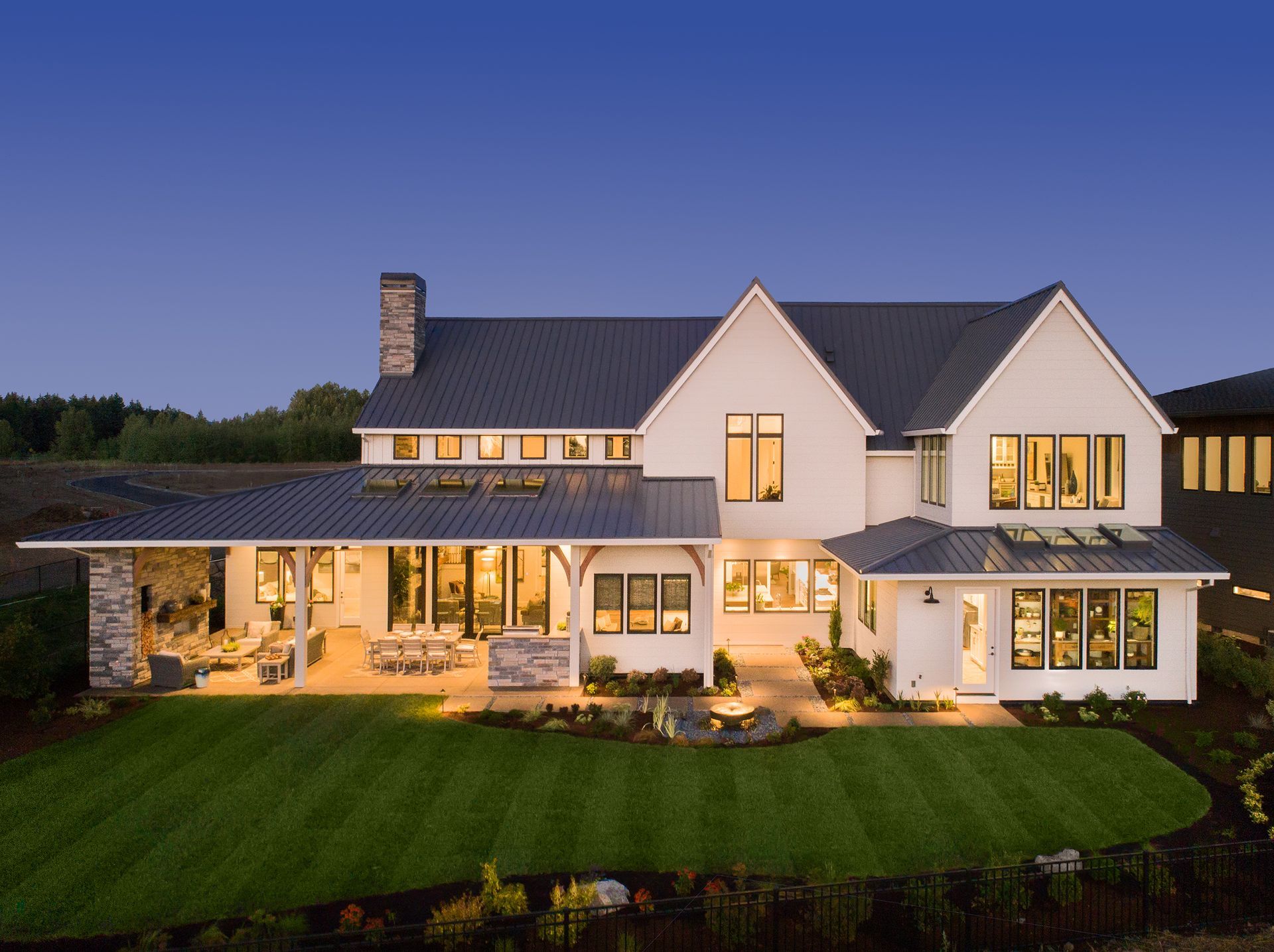 White farmhouse with dark roof and large porch, illuminated at dusk, green lawn.
