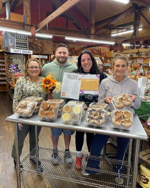 Four smiling people stand behind a metal table display of various baked goods in a rustic indoor market.