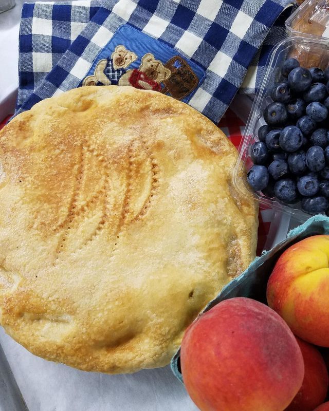 A golden-brown pie with decorative leaf imprints, sits next to a container of blueberries and fresh peaches on a cloth.