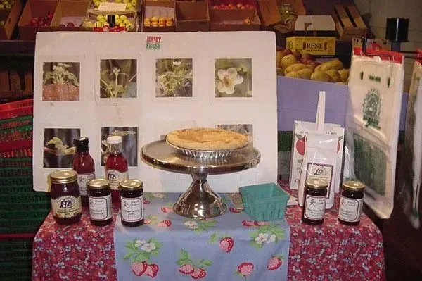 A display of fruit products and a pie on a stand, set on a strawberry-print table cloth in front of a botanical poster.