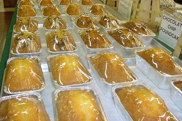 Rows of individually wrapped golden loaf cakes and muffins displayed in aluminum pans at a bakery or market.