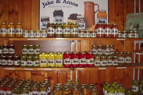 Wooden shelves stocked with numerous glass jars of Jake & Amos brand food products against a wood-paneled wall.