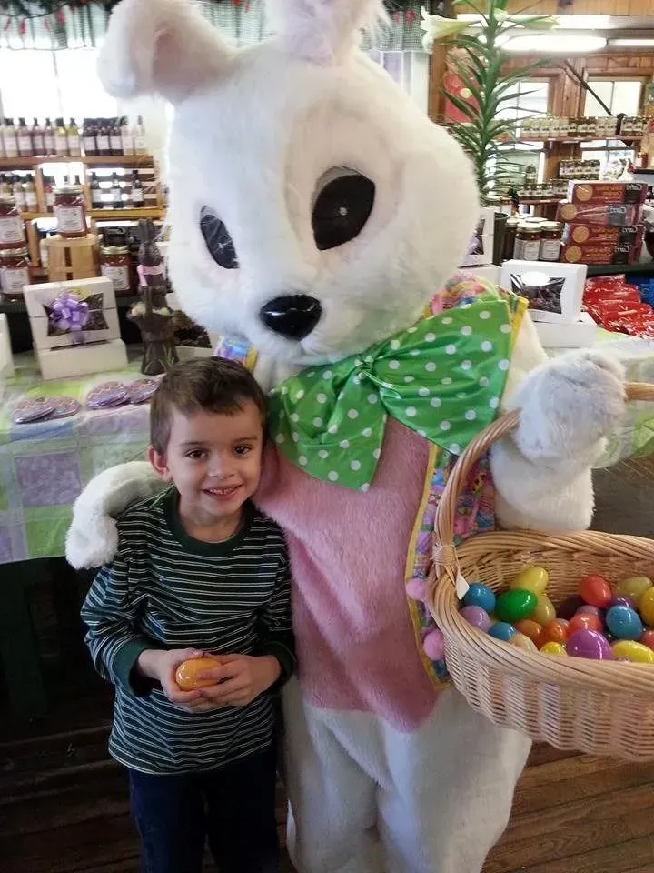 A child smiles while standing next to a person in a white Easter bunny costume holding a basket of colorful plastic eggs.
