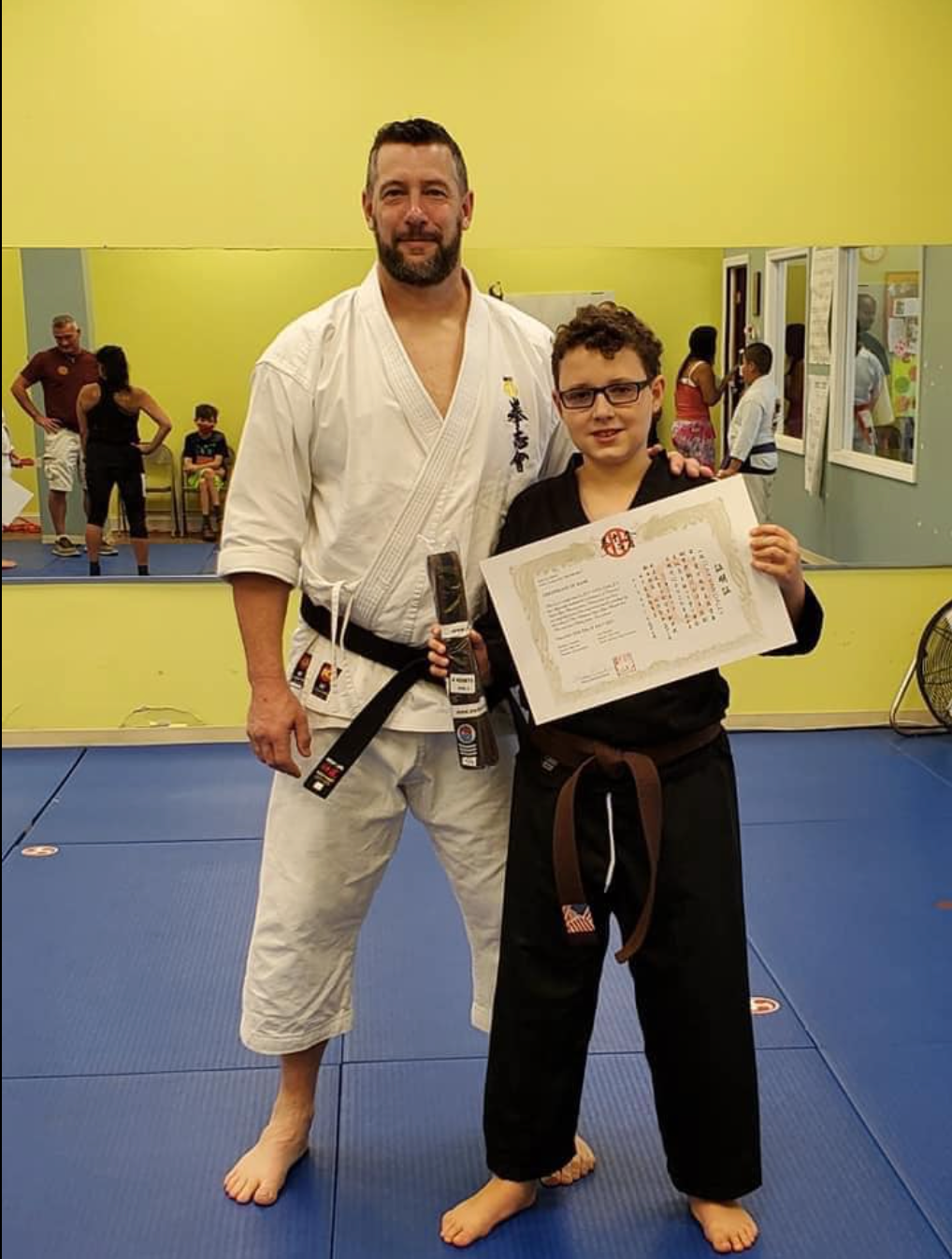 A man in a white karate uniform is standing next to a boy in a black belt holding a certificate.