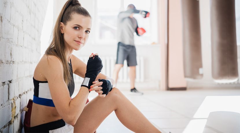 A woman is sitting on the floor in a gym with boxing gloves on her hands.
