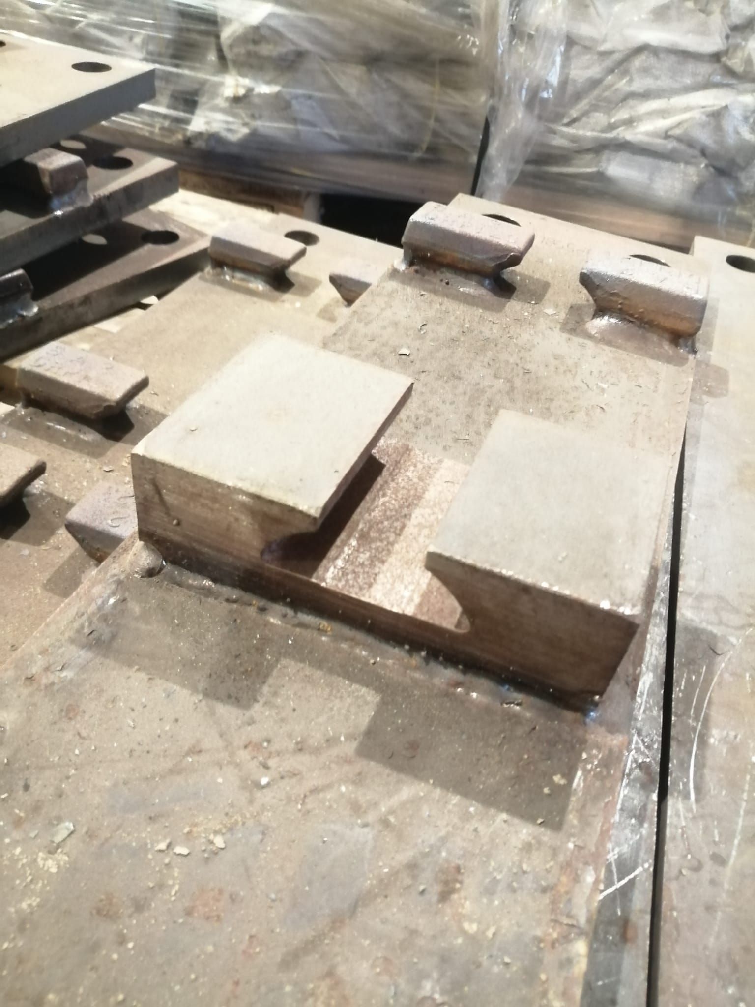 Una pila di grandi lastre metalliche industriali grigie con blocchi di montaggio sporgenti e scanalati, appoggiate sul pavimento di un'officina.