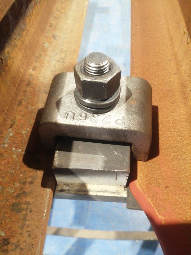 Un morsetto metallico per travi, fissato con un bullone e una rondella, su un blocco metallico rettangolare posto su una rotaia industriale in acciaio arrugginita.
