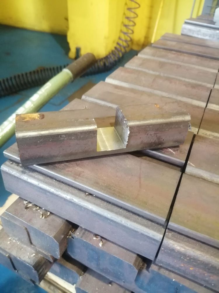 Un tubo metallico a sezione quadrata con una tacca, appoggiato su una pila di pezzi identici in un'officina.