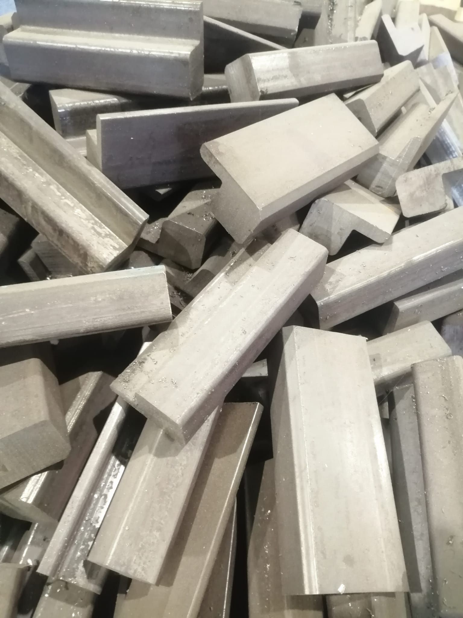 Un mucchio di elementi strutturali metallici a forma di L, di colore grigio chiaro, accatastati in modo disordinato.