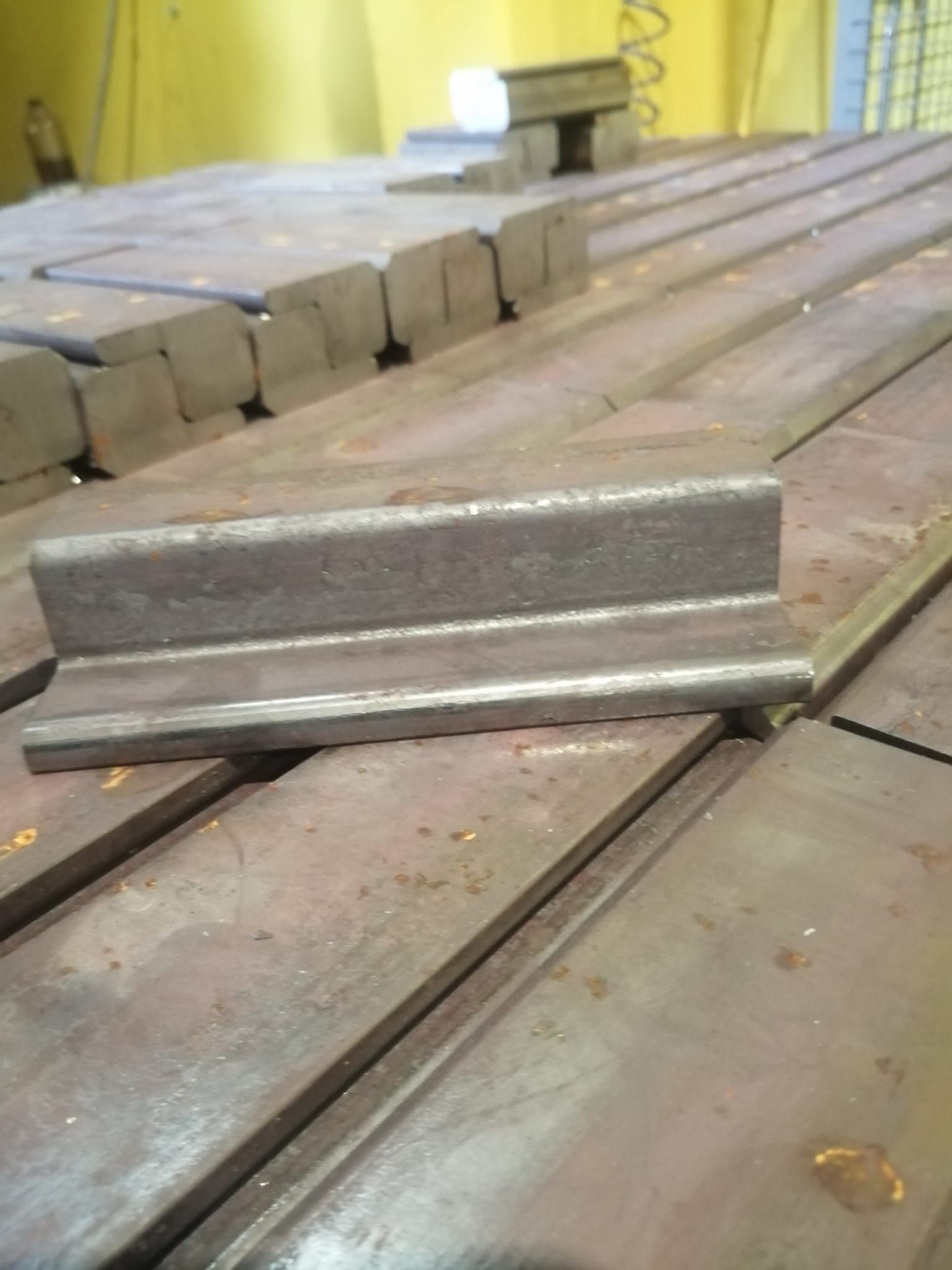 Una piccola staffa angolare in metallo grigio è appoggiata su una superficie di lavoro industriale piana e scanalata, tra pezzi di metallo simili.