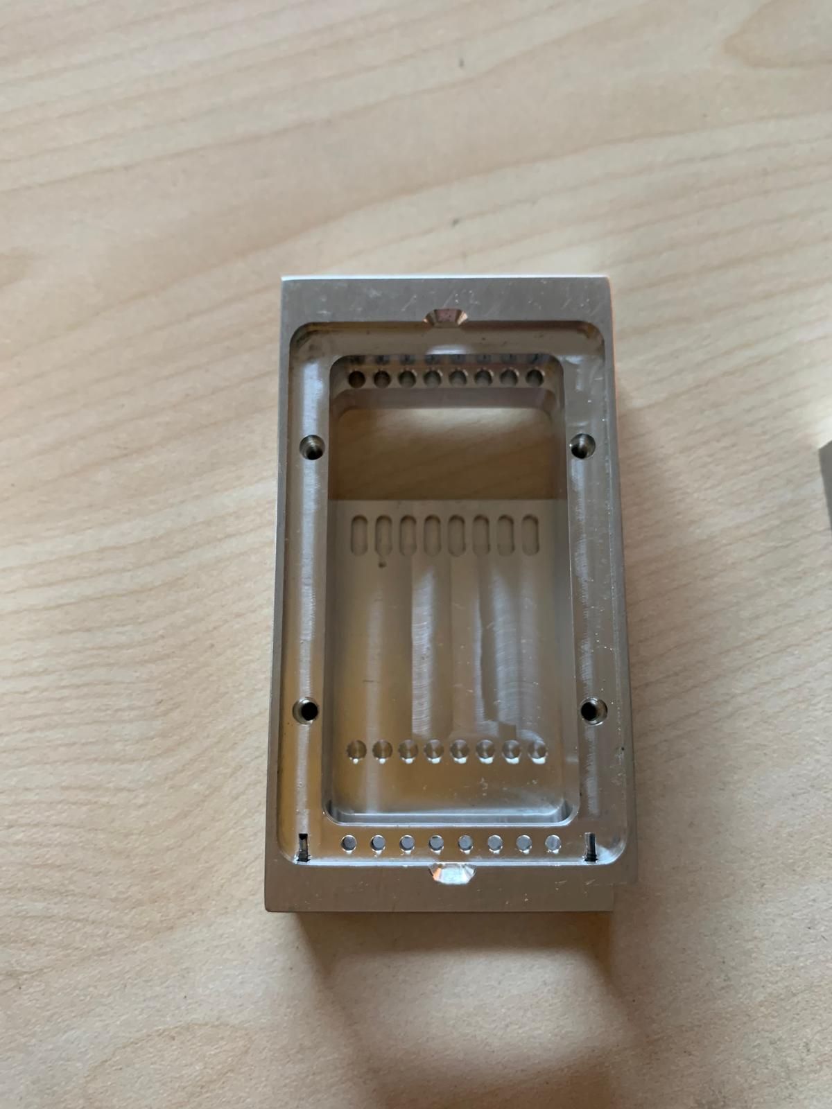 Un alloggiamento rettangolare in metallo lavorato, di colore argento, con fessure interne e fori di montaggio su una superficie in legno.