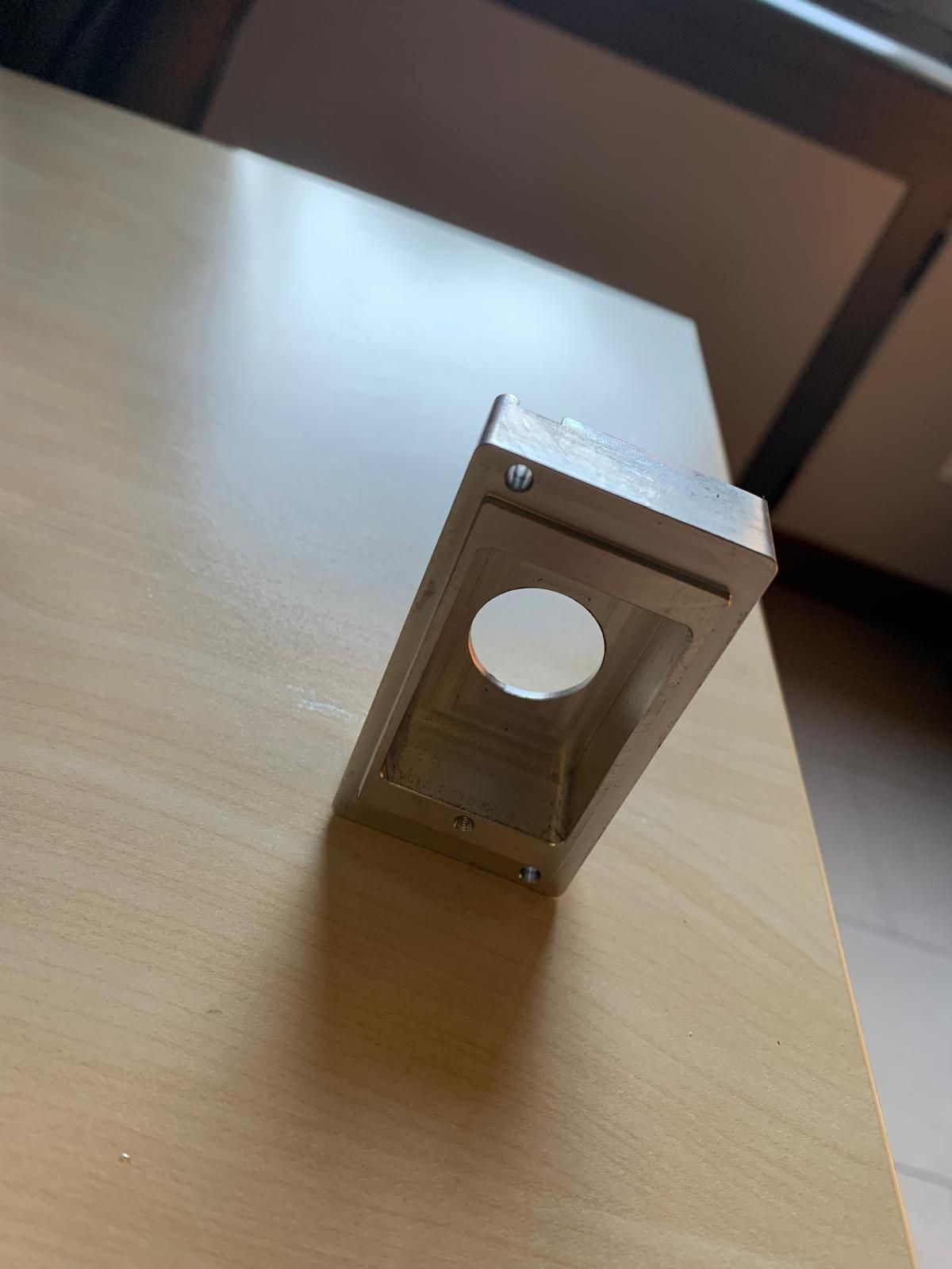 Una staffa metallica rettangolare con un foro circolare, centrata su una superficie di legno chiaro.