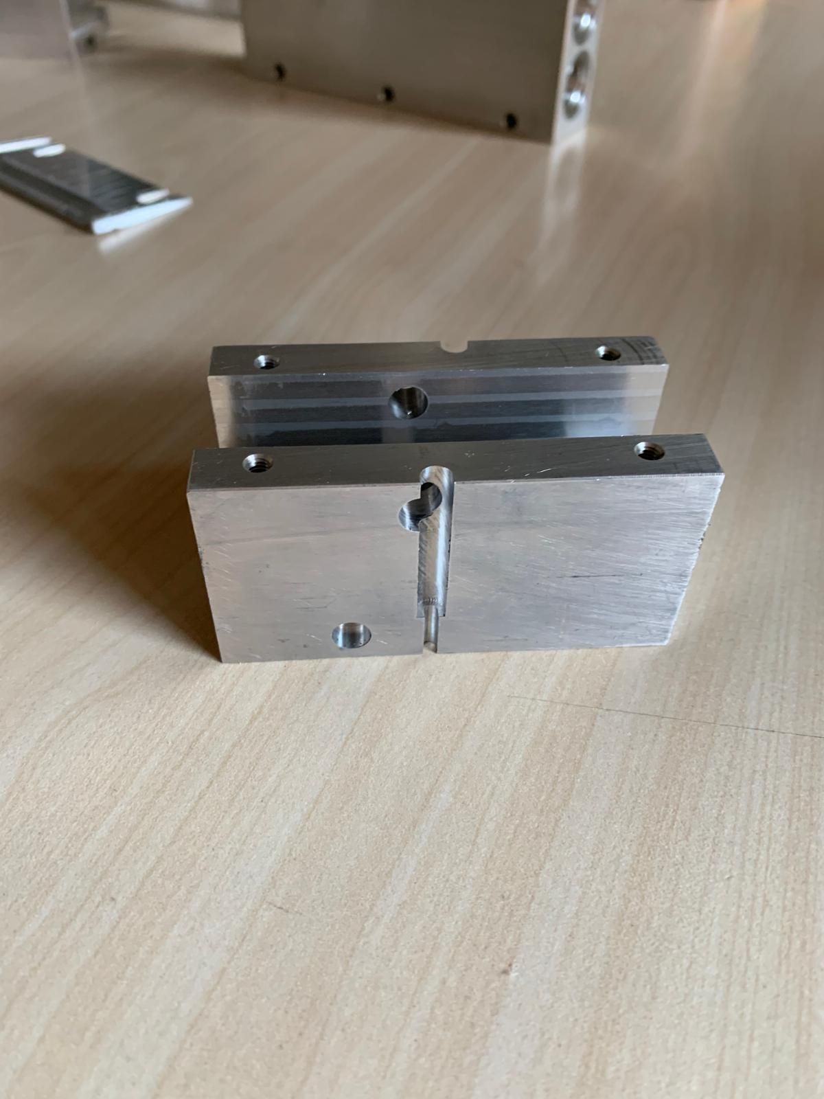 Due elementi rettangolari in alluminio lavorato, di colore argento e con fori preforati, poggiano su un tavolo con venature in legno chiaro.