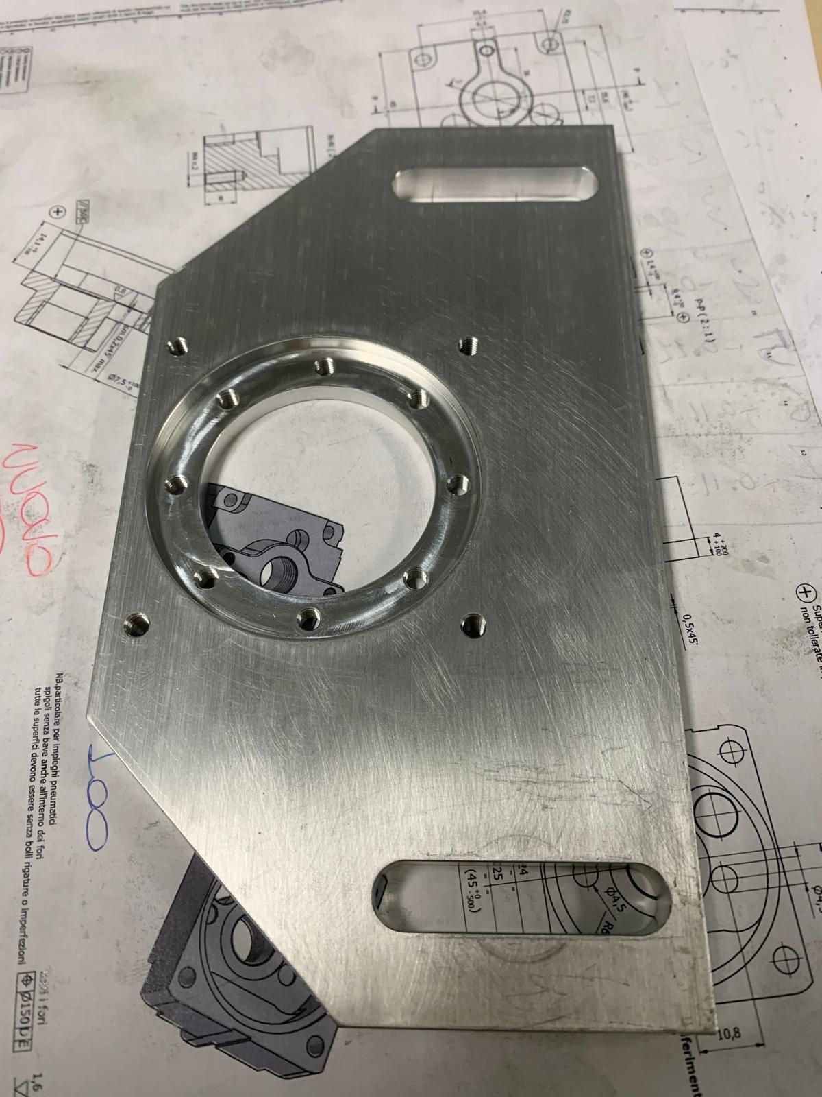 Una piastra metallica argentata lavorata con un foro circolare centrale e fori di montaggio, appoggiata su un disegno tecnico.