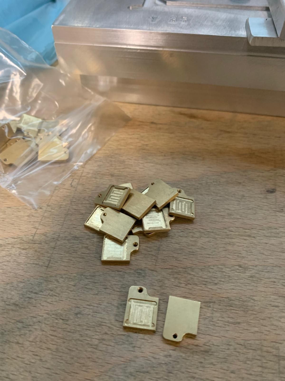 Piccole targhette metalliche rettangolari color oro con motivi incisi e fori, sparse su un banco da lavoro in legno vicino a un attrezzo.