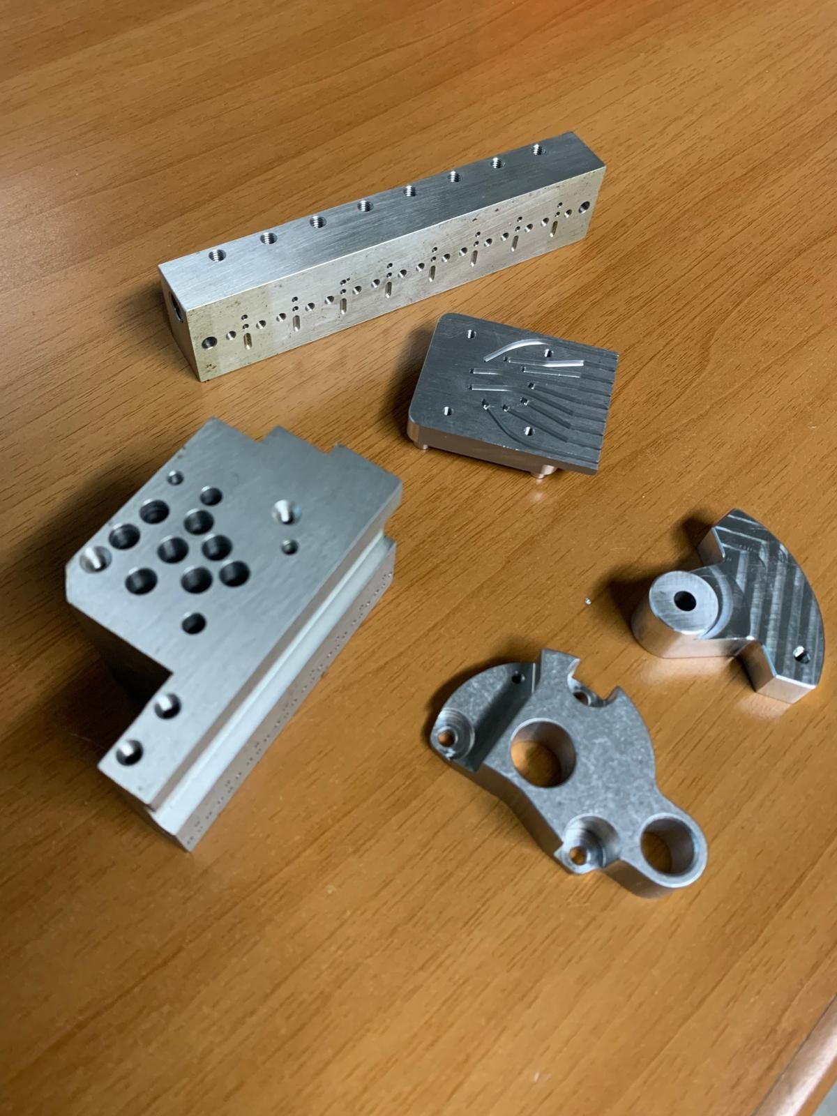 Cinque componenti metallici lavorati con fori e intricate lavorazioni di fresatura, esposti su una superficie di legno.