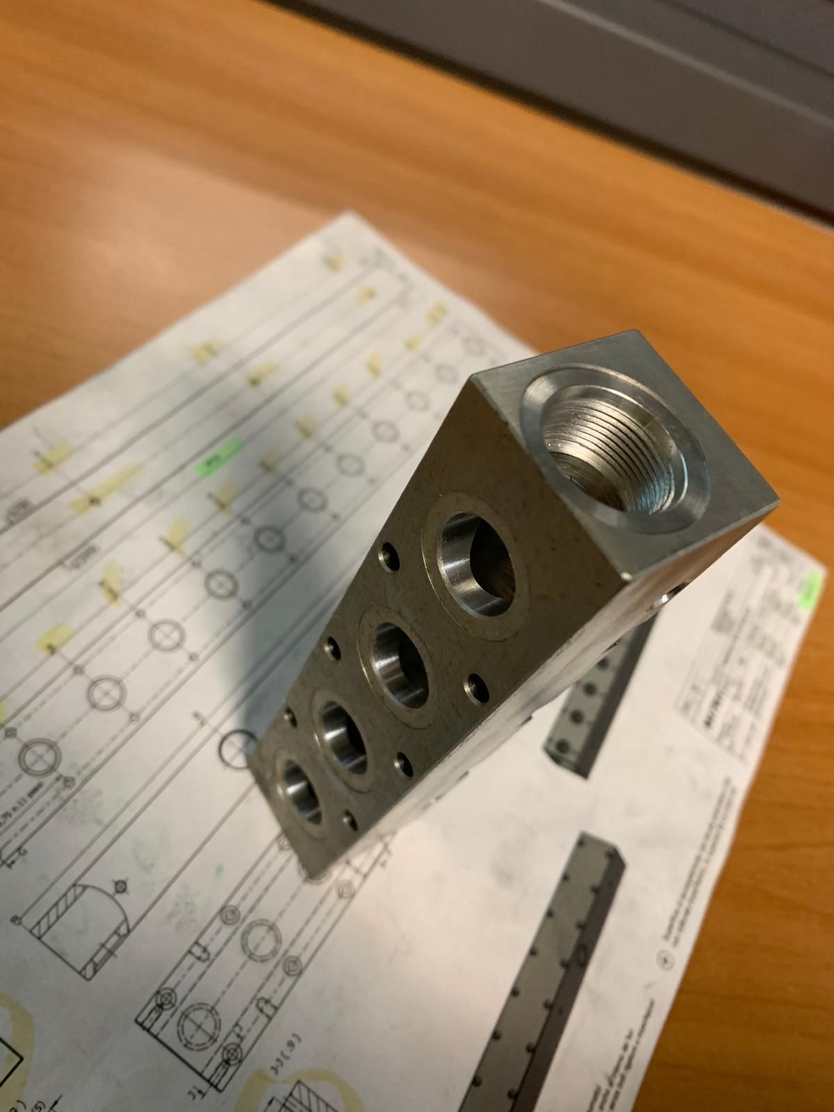 Un blocco collettore metallico rettangolare con porte filettate, appoggiato su disegni tecnici su una scrivania di legno.