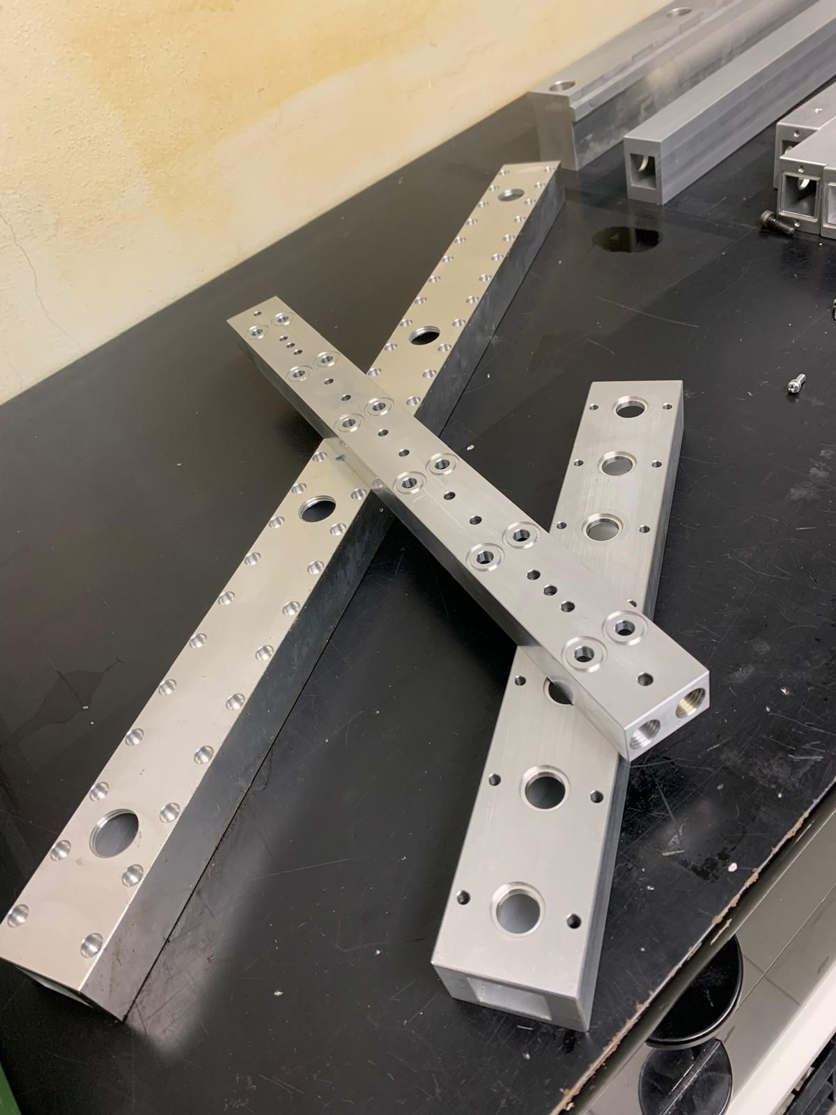 Tre profilati di alluminio lavorati, con fori preforati e punti di fissaggio, su un banco da lavoro nero.