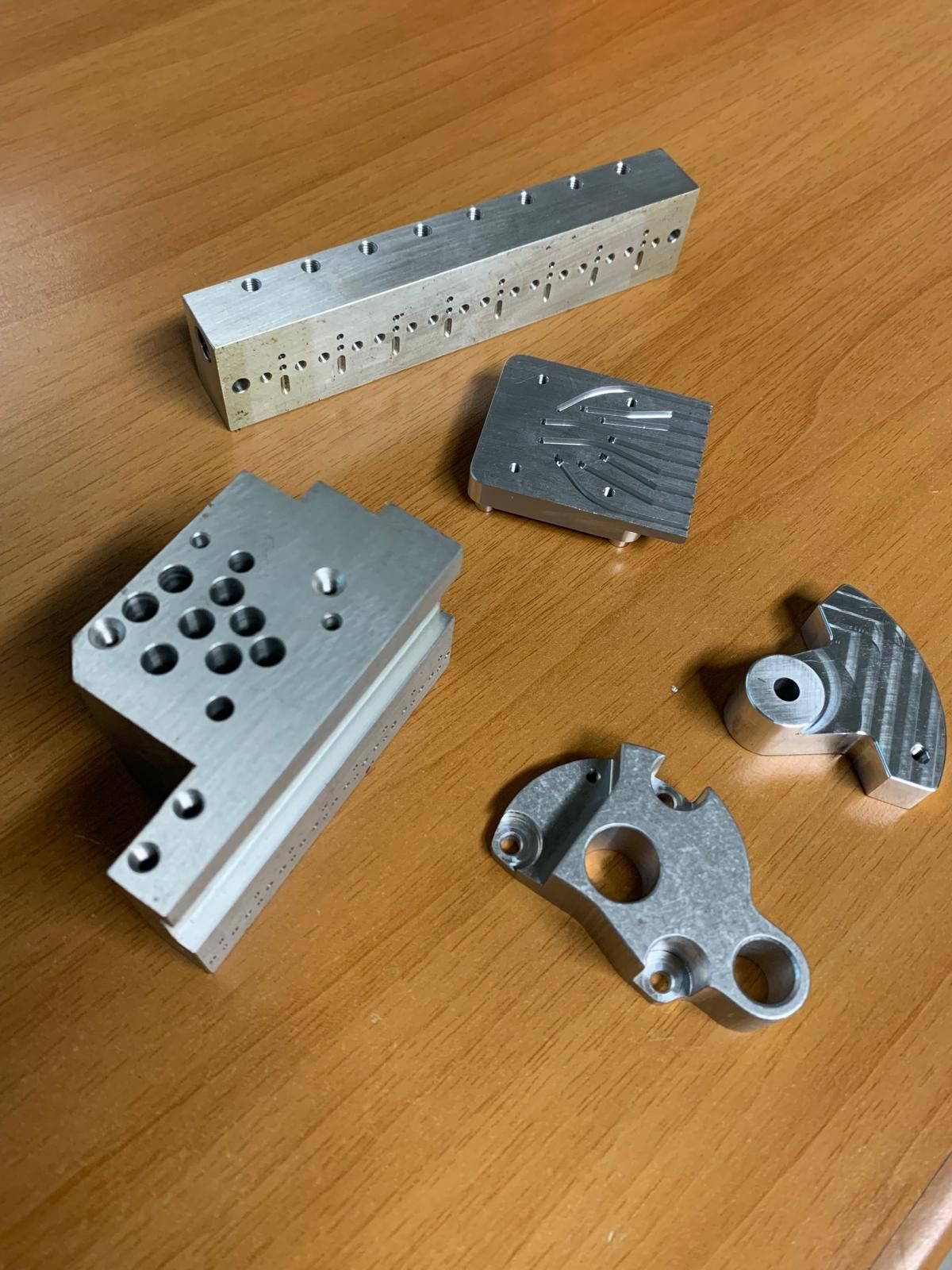 Diversi componenti in alluminio lavorato, tra cui un lungo blocco rettangolare e varie staffe, poggiano su una superficie di legno.