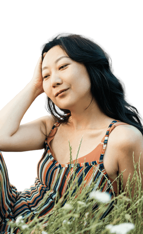 Woman in striped jumpsuit, hand in hair, sitting in tall grass, looking relaxed.