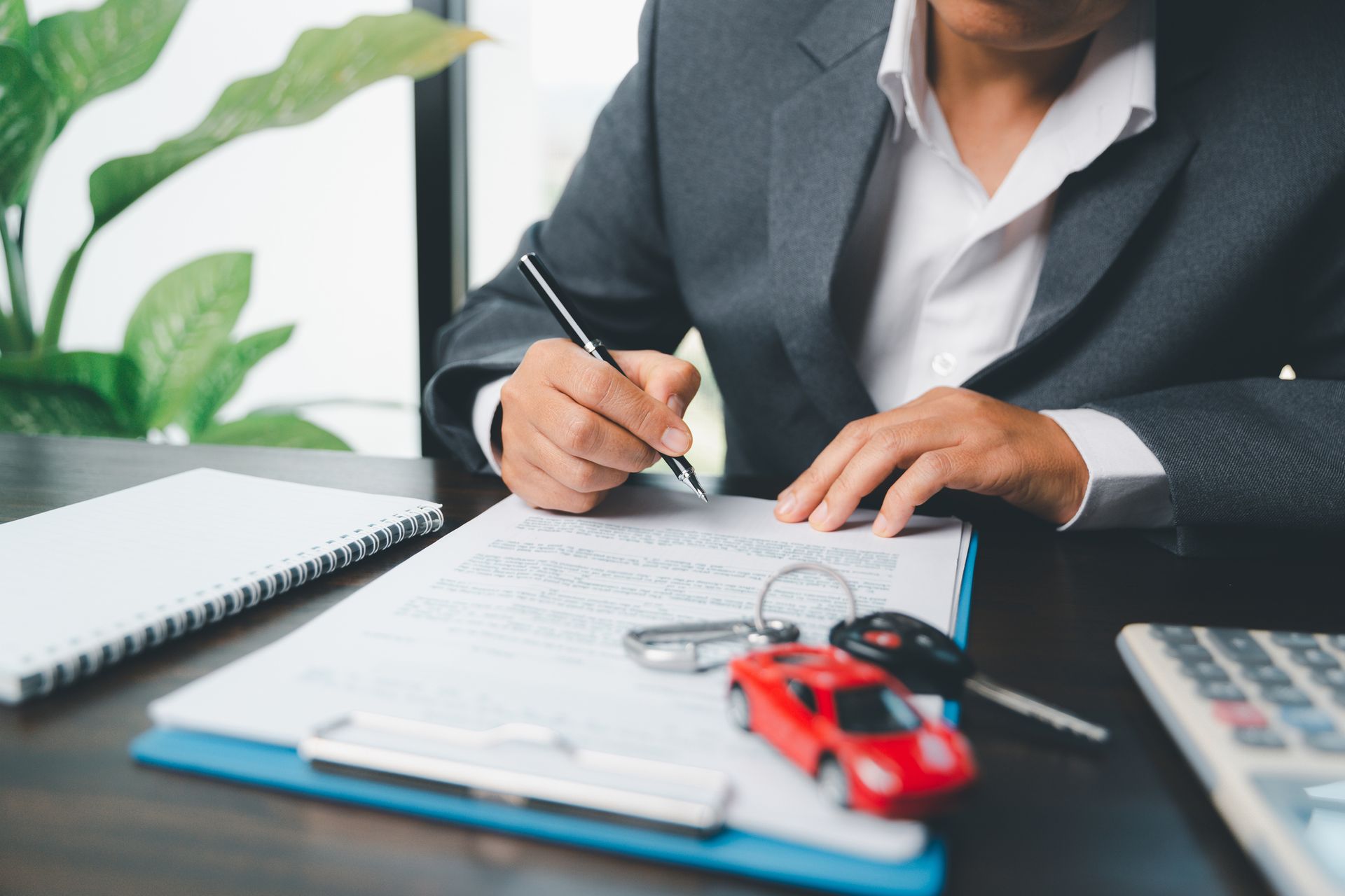 A man in a suit is signing a car protection insurance document, seated at a desk with his car keys.