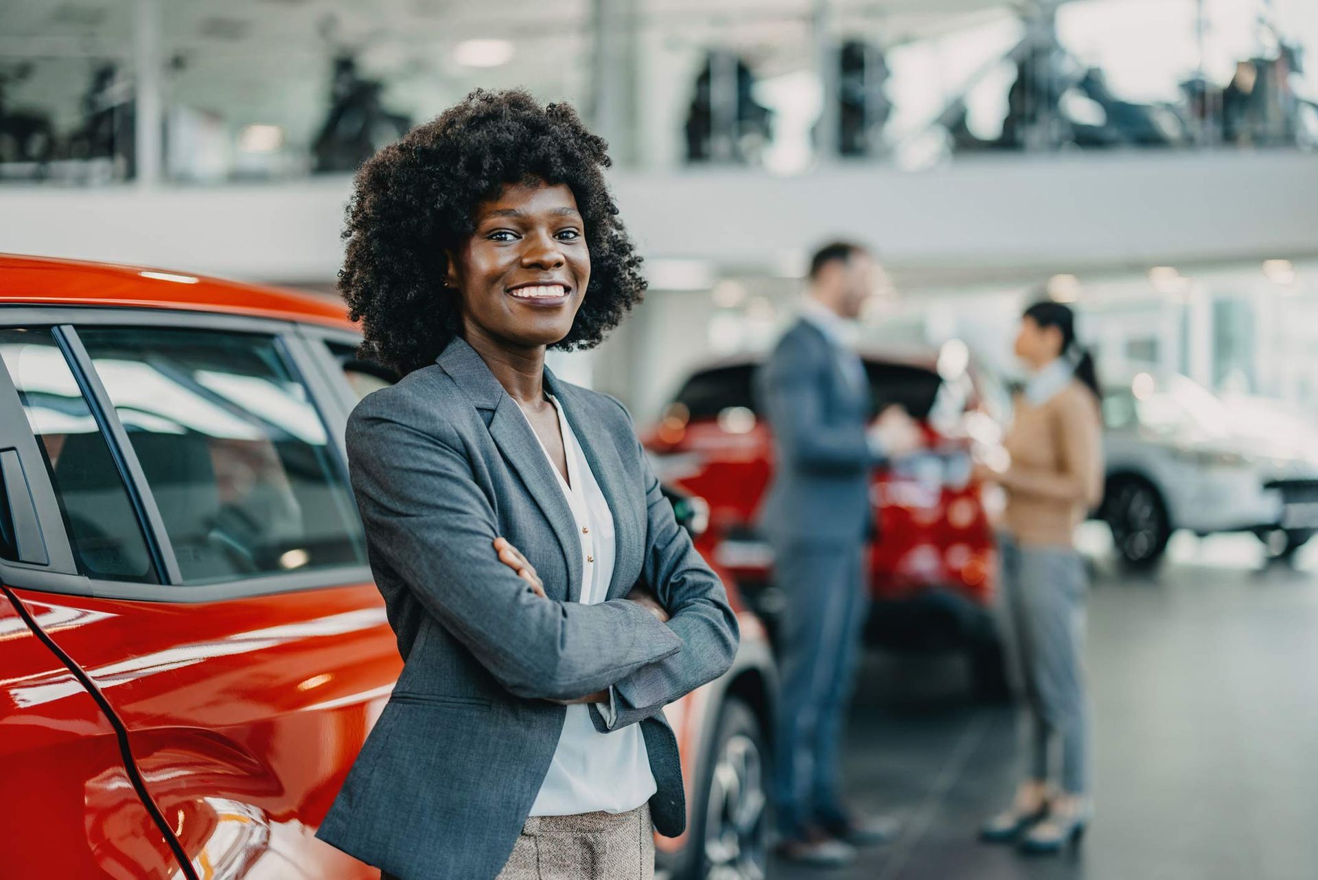 Woman in a car showroom with arms crossed with two people in the background.