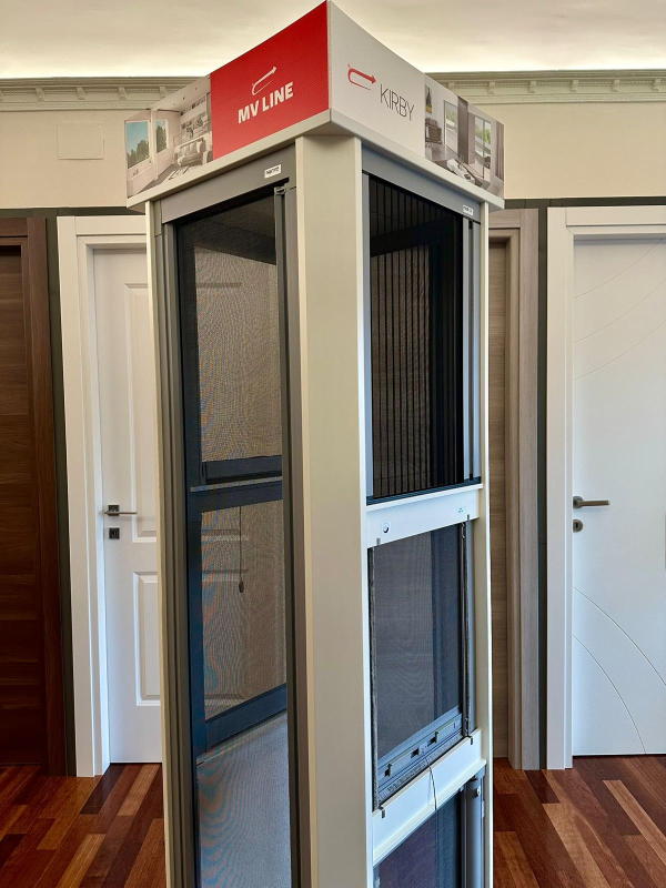 Cabina espositiva bianca per ascensori con insegne rosse in una stanza con pavimento e porte in legno sullo sfondo.