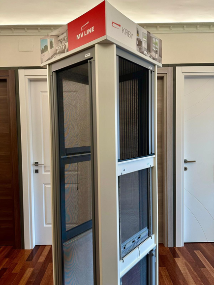 Cabina espositiva bianca per ascensori con insegne rosse in una stanza con pavimento e porte in legno sullo sfondo.
