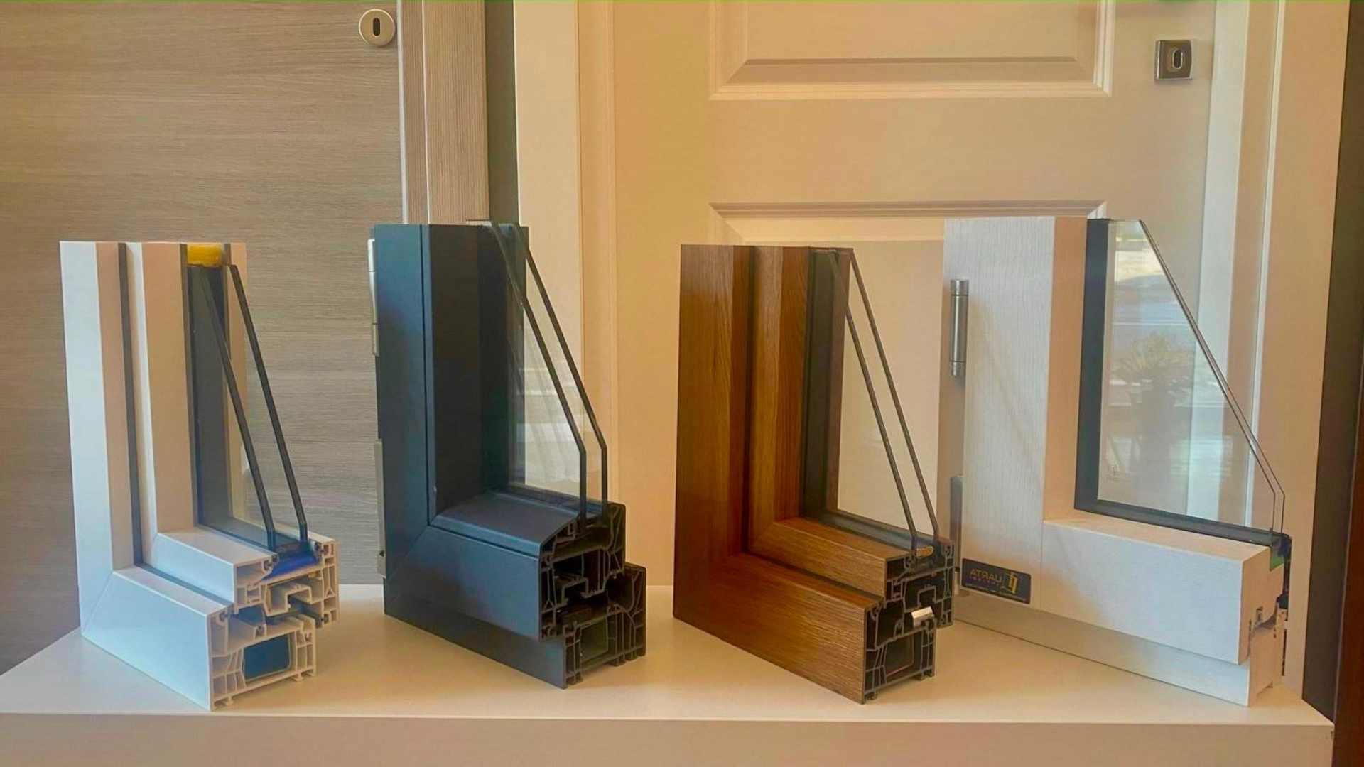 Tre campioni di telai per finestre moderni, in bianco, legno e nero, esposti su un tavolo all'interno.