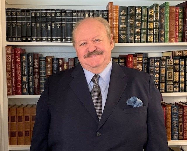 A person in a dark suit, tie, and blue pocket square smiles in front of a bookshelf filled with many books.