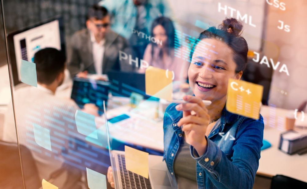 A woman is holding a laptop and writing on a glass wall.