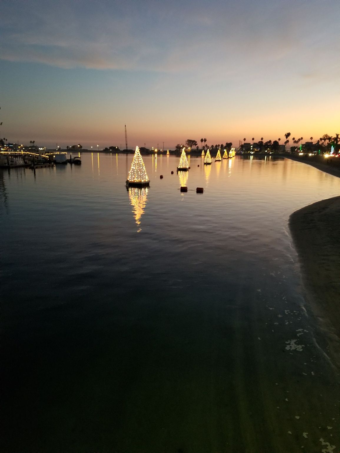 Bay with Christmas tree lights at dusk. Water reflects soft sunset colors; shoreline to the right.