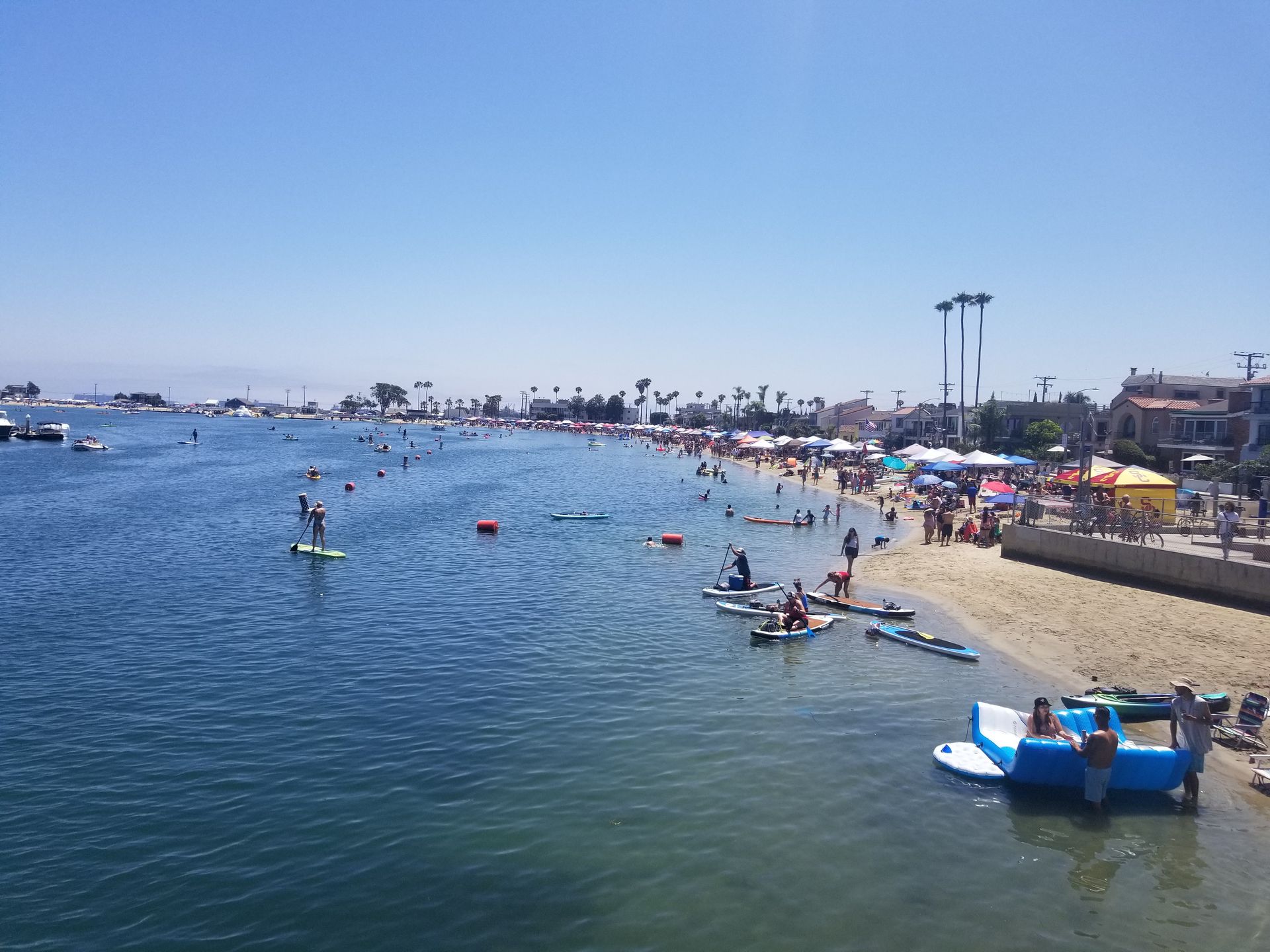 Beach scene with people swimming, paddleboarding, and relaxing on the sand under a blue sky.