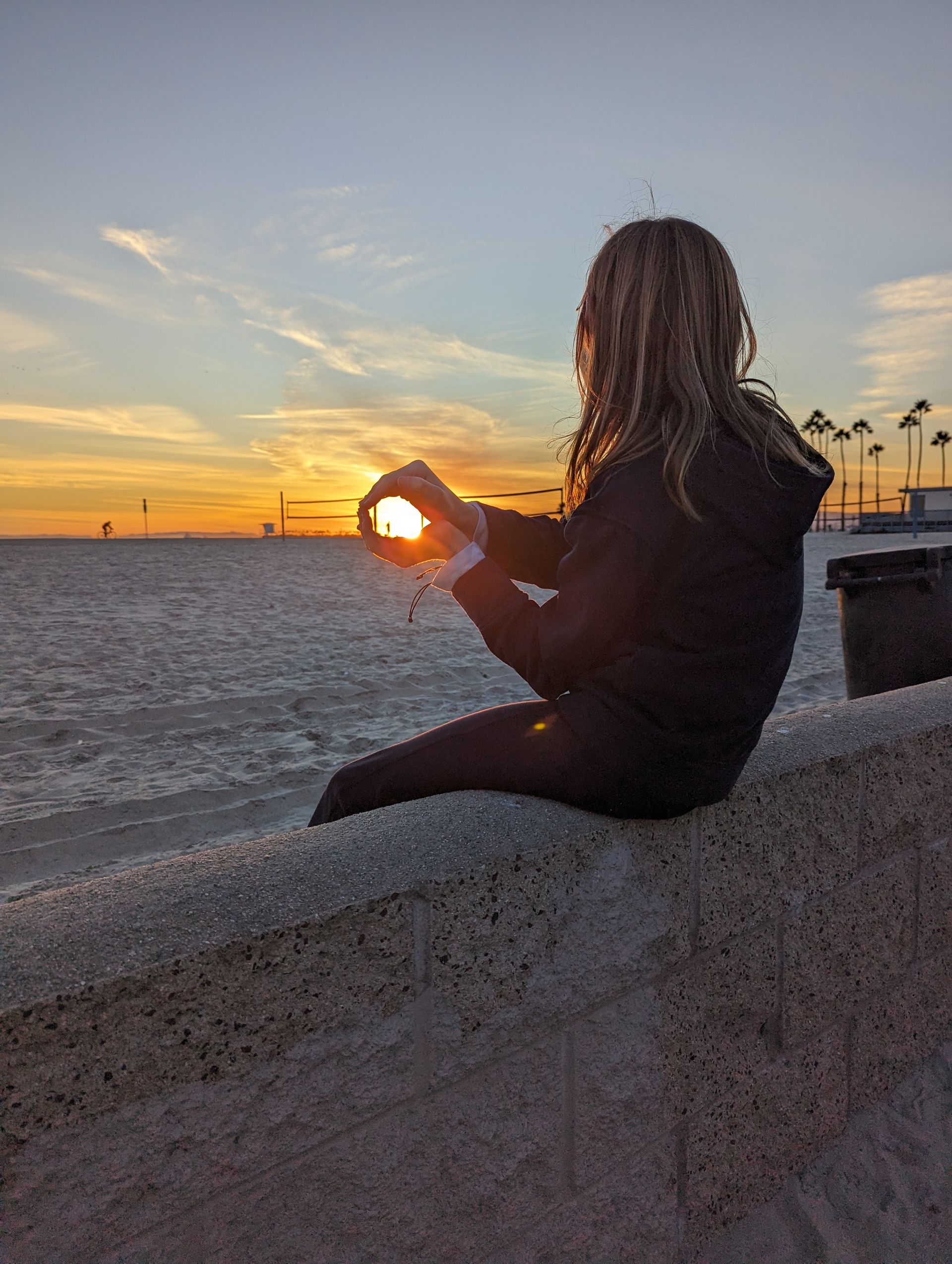 Person holding up the sun, silhouetted against a sunset on a beach.