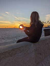 Person sitting on a wall holding the setting sun, silhouetted against an orange and blue sky. Beach background.