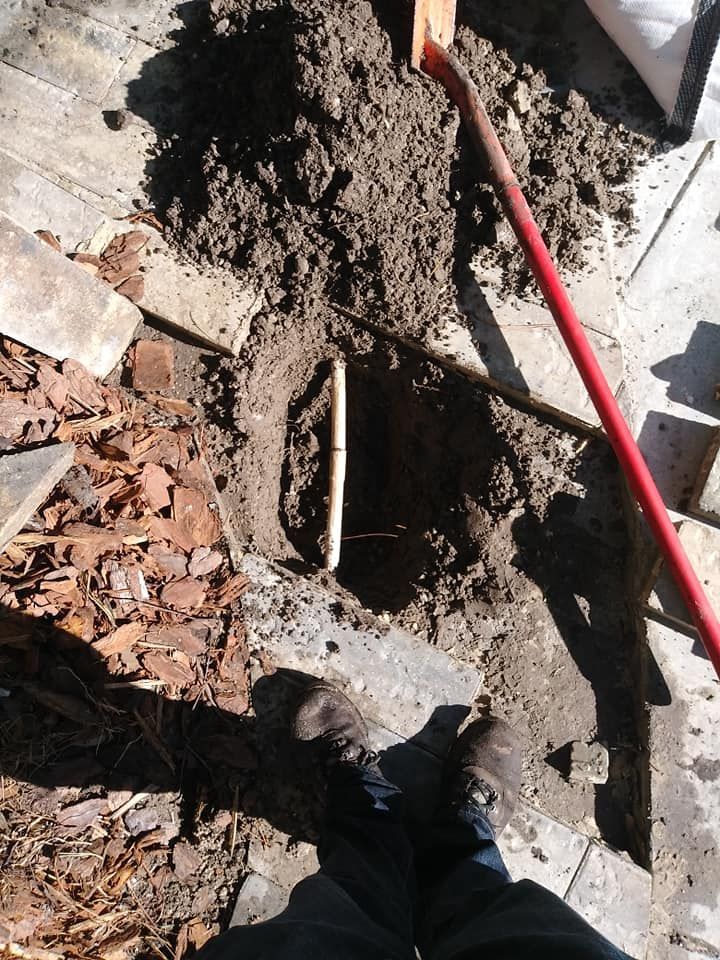 Hole dug in patio, revealing a white pipe. A red handled tool is leaning into the hole; feet are visible.