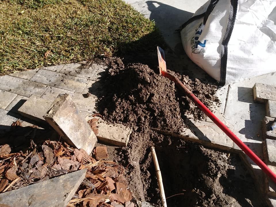 Shovel digging in dirt next to paving stones and landscaping, with a bag in the background.