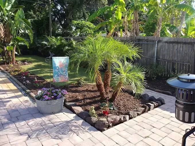 Palm tree in a landscaped garden with brick pavers, a decorative flag, and a metal container with flowers.