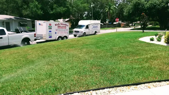 White truck towing a trailer and a white van parked on a green lawn, street in the background.