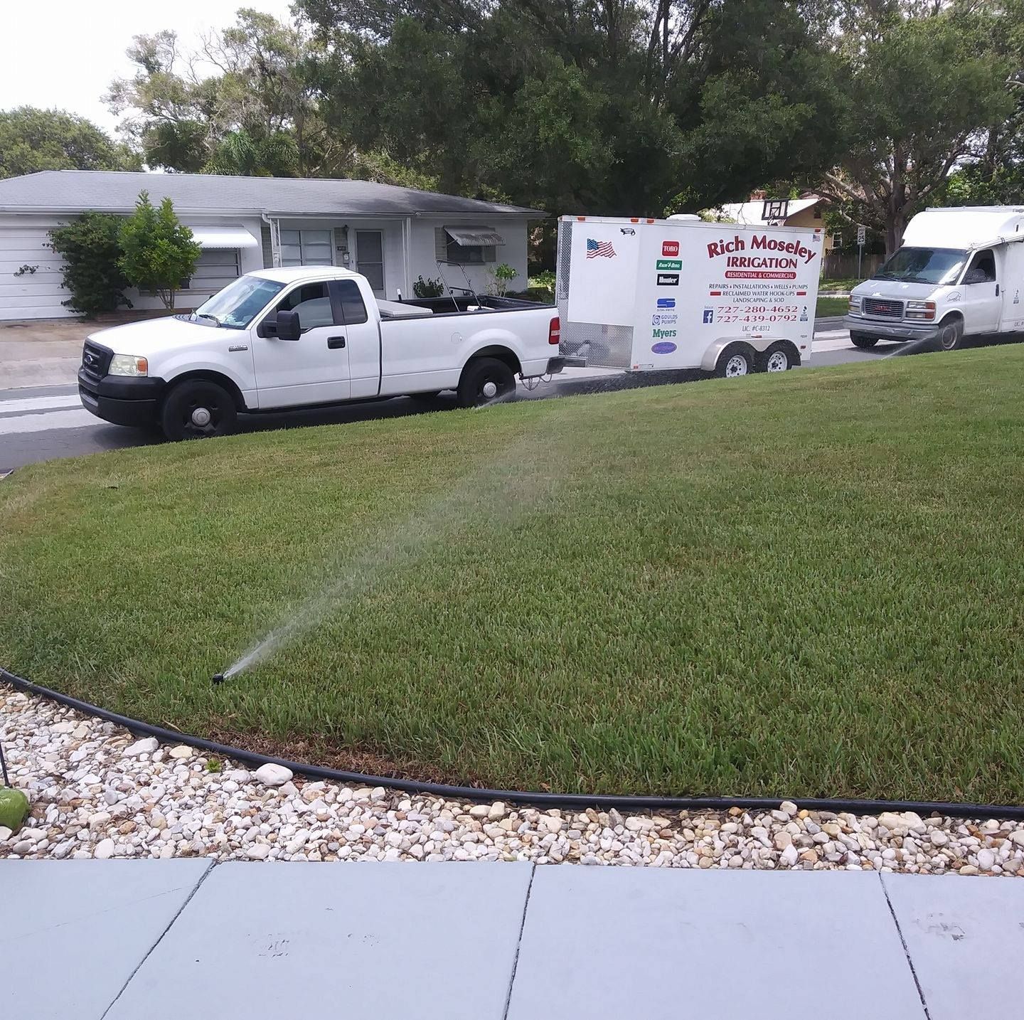 A white pickup truck pulling a trailer, and a white van parked on grass with a sprinkler running.