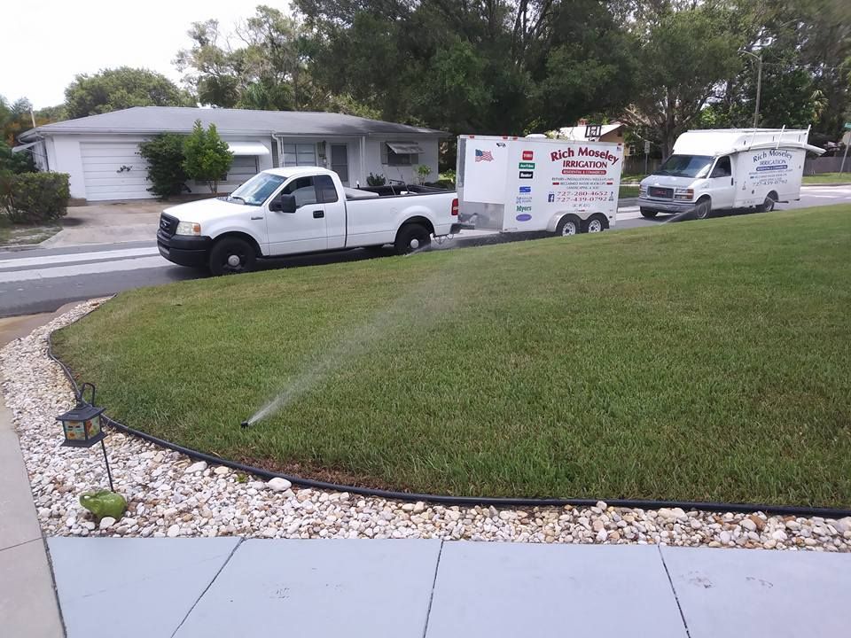 White pickup truck towing a trailer and a white van parked on a suburban lawn with a sprinkler running.