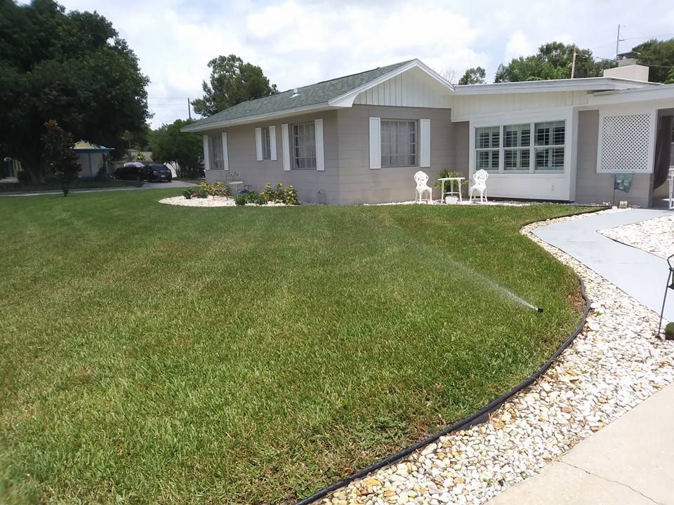 Lawn being watered in front of a light gray house with white trim. White rocks and walkway. Green grass and trees.
