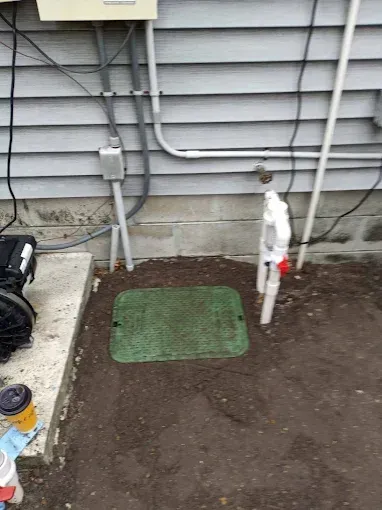 Green irrigation valve box on dirt next to a house with electrical and water pipes.
