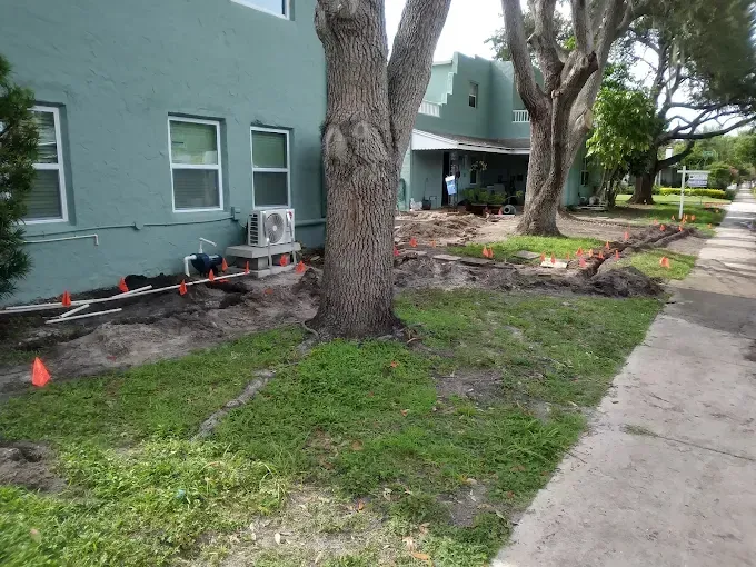 Excavated landscaping area with orange markers near a light green building and trees, next to a sidewalk.