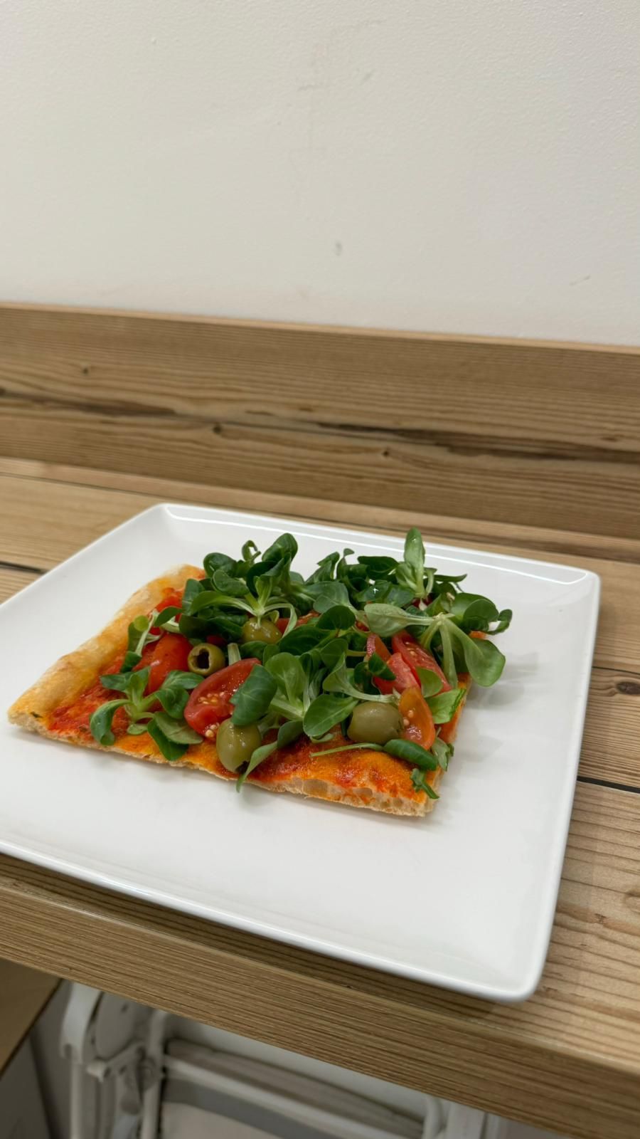 Una fetta di pizza con pomodori, olive e lattuga su un piatto bianco su un tavolo di legno.